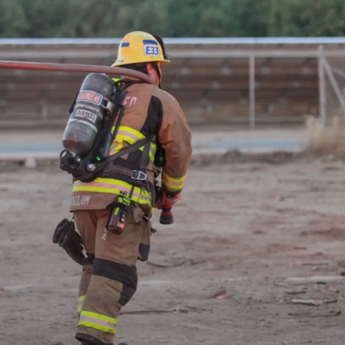 A firefighter in full turnout gear walks through an outdoor training area. The firefighter is wearing a yellow helmet, tan fire protective suit with reflective yellow stripes, SFL radio harness and a large oxygen tank on their back.