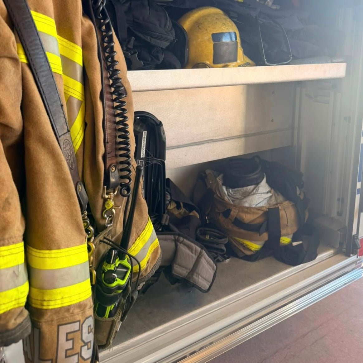 A firefighter turnout jacket with reflective yellow stripes hangs on the left side of an open storage compartment, with other firefighting gear including a helmet, straps, and bags stored inside.