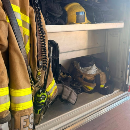 A firefighter turnout jacket with reflective yellow stripes hangs on the left side of an open storage compartment, with other firefighting gear including a helmet, straps, and bags stored inside.