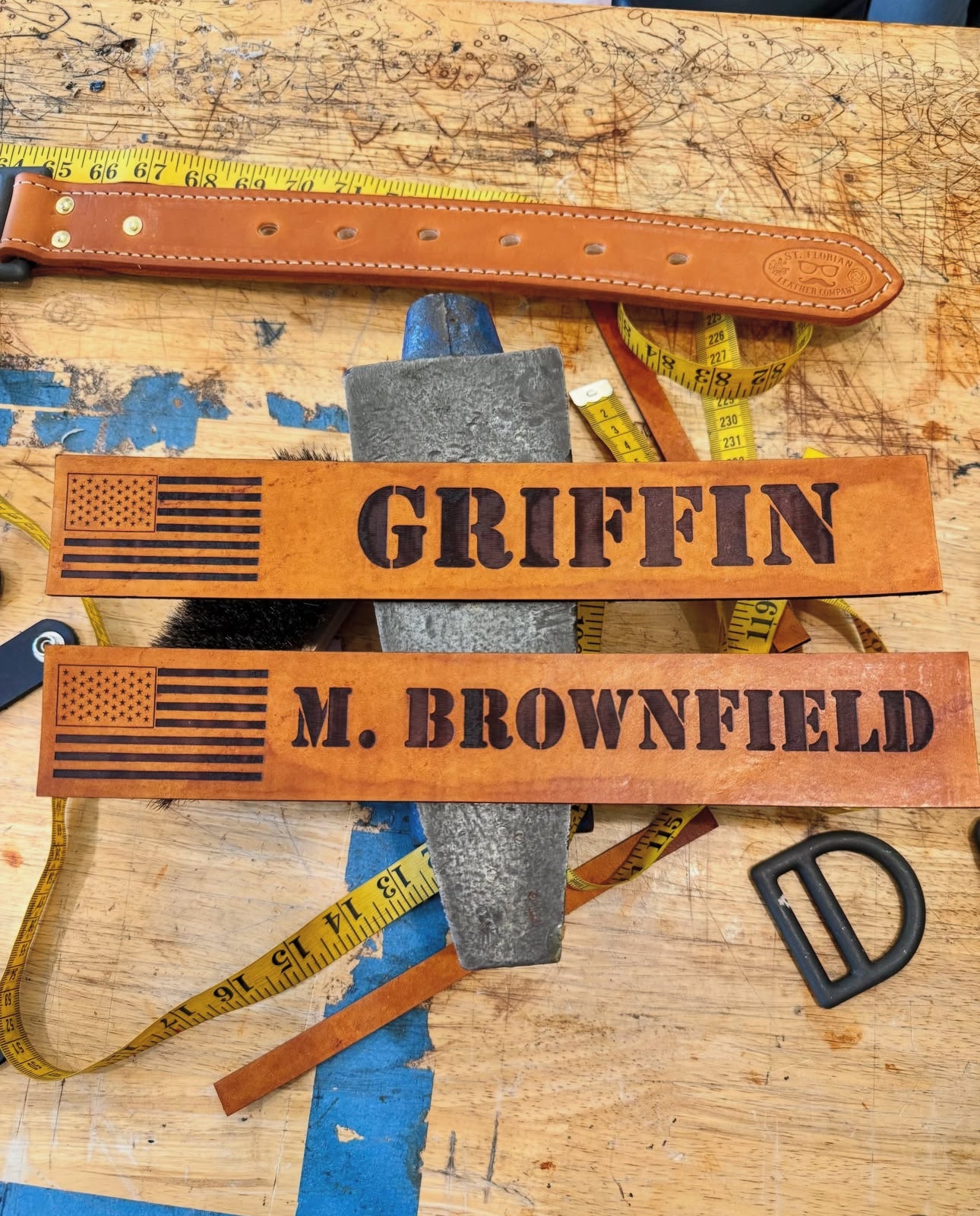 Two personalized brown leather nameplates engraved with 'GRIFFIN' and 'M. BROWNFIELD' alongside a black American flag design, displayed on a workbench with a measuring tape, hammer, and metal hardware.