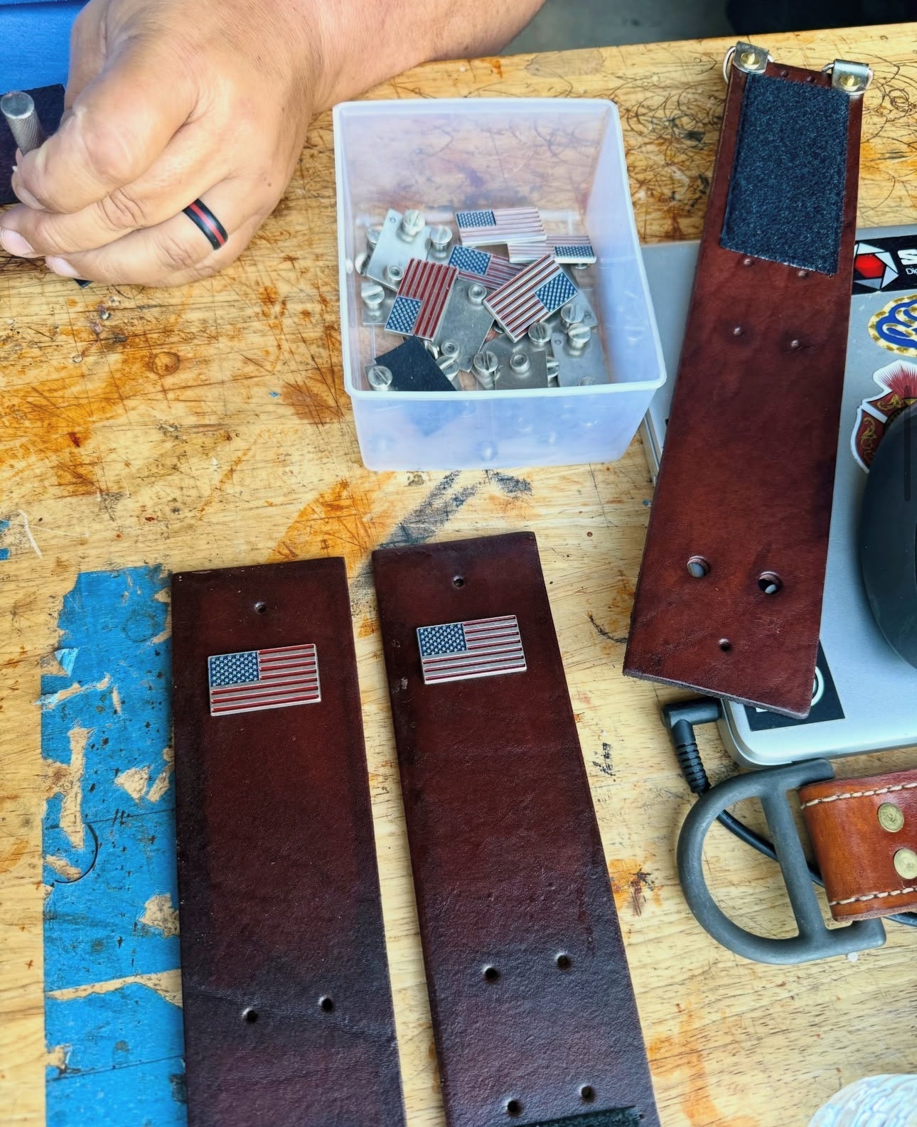 Two dark brown leather belt radio harnesses with small American flag pins, laid out on a wooden workbench next to a hand, a container of metal hardware, and other crafting materials.
